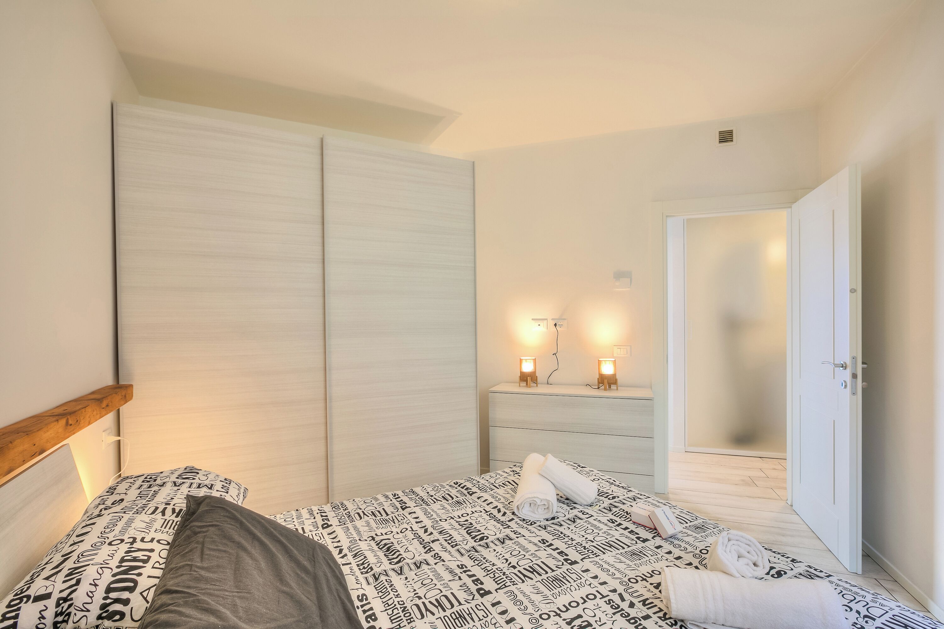 Ein Schlafzimmer mit einem Bett, einem Nachttisch mit zwei Wandlampen, einem Kleiderschrank und einer Tür zu einem anderen Raum.