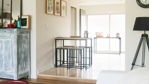 A modern living room with a white sofa, a coffee table, and a dining area with a table and chairs. There is a large window providing natural light and a view of the outside.
