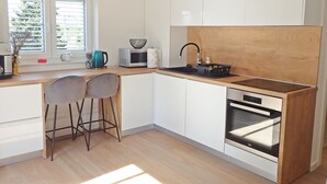 Een moderne keuken met witte kasten, een houten aanrecht, een spoelbak, een waterkoker, een magnetron en een inbouwoven. Er staan twee barkrukken en er is een raam met jaloezieën.