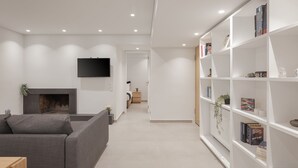 Uma sala de estar moderna com um sofá cinza, uma televisão de tela plana na parede, uma lareira e uma estante repleta de livros e objetos decorativos.