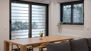 Uma área de refeições com mesa e cadeiras de madeira, janela com vista para o verde e um pequeno vaso com flores no parapeito.