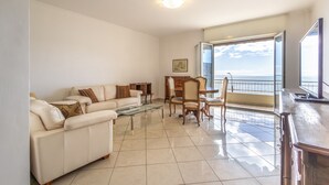 A living room with a sofa, coffee table, and dining area with chairs. There is a large window with a view of the sea.