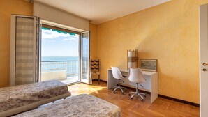 A bedroom with two beds, a desk with chairs, a window with a view of the sea, and a door.