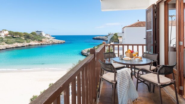 A balcony with a table set for breakfast overlooking a turquoise sea and a sandy beach.