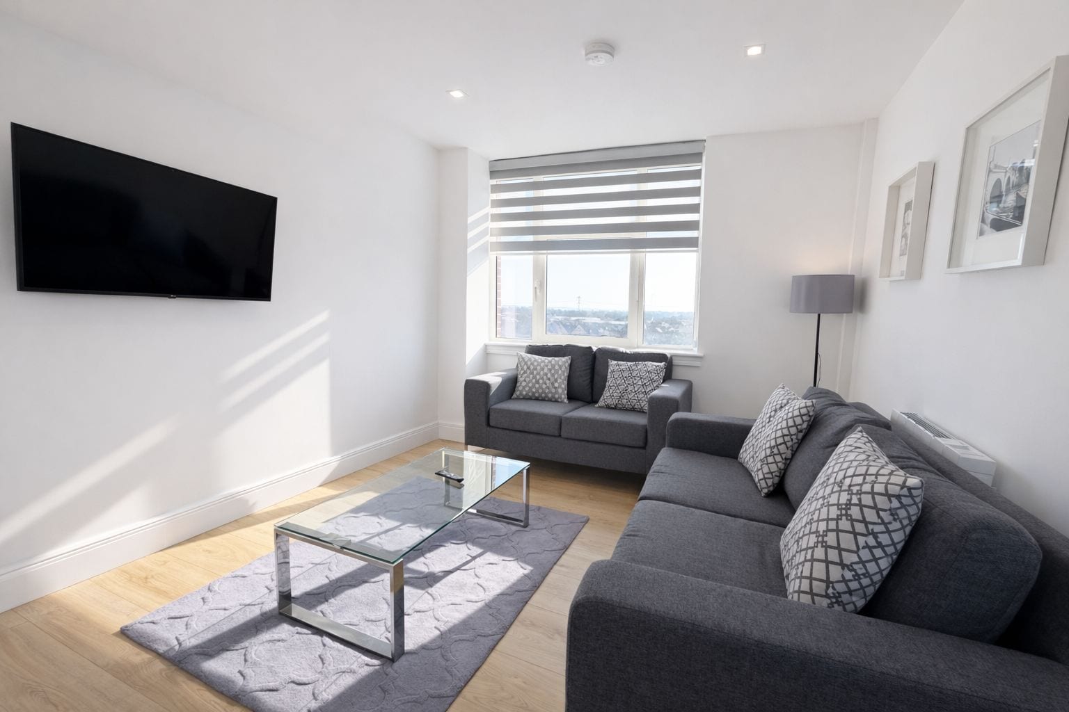 A modern living room with a grey sofa, a glass coffee table, and a flat-screen TV mounted on the wall.