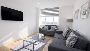 A modern living room with a grey sofa, a glass coffee table, and a flat-screen TV mounted on the wall.