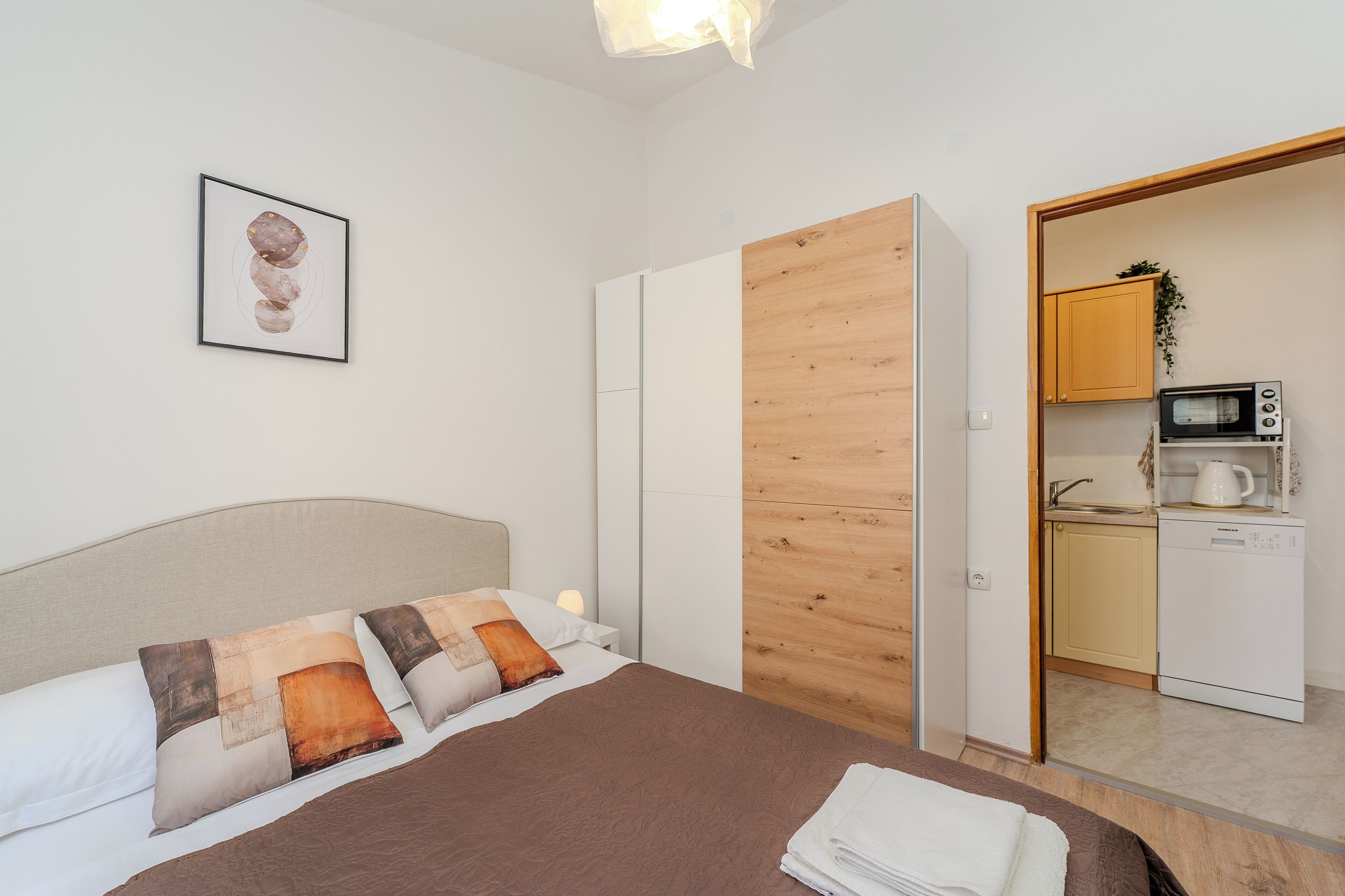 A bedroom with a bed, pillows, a nightstand, a framed picture, a wooden wardrobe, and a kitchenette with a microwave, kettle, and sink.
