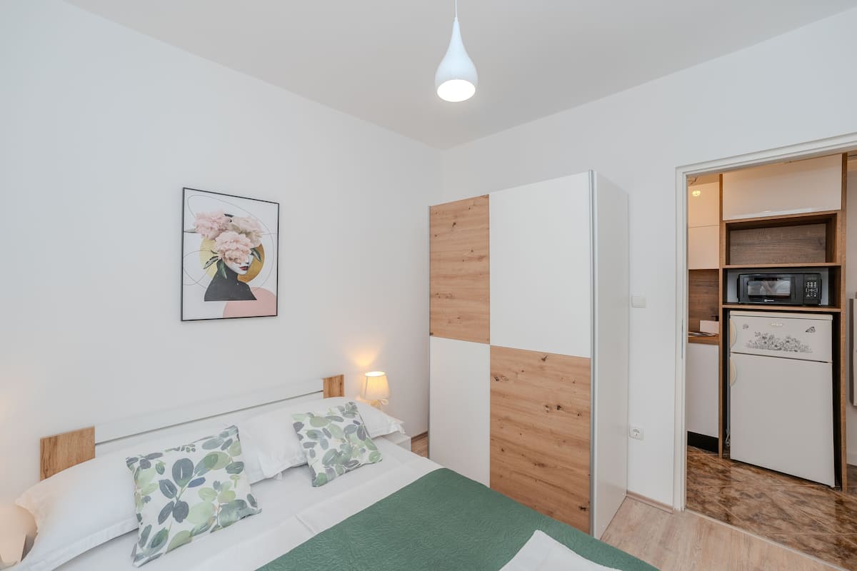 A bedroom with a bed, pillows, a nightstand, a lamp, a wooden wardrobe, and a microwave in the adjacent kitchen.