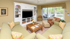 Ein Wohnzimmer mit Fernseher, Bücherregalen, zwei Sofas mit floralen Kissen, einem Korbtisch und einer Glastür zum Garten.