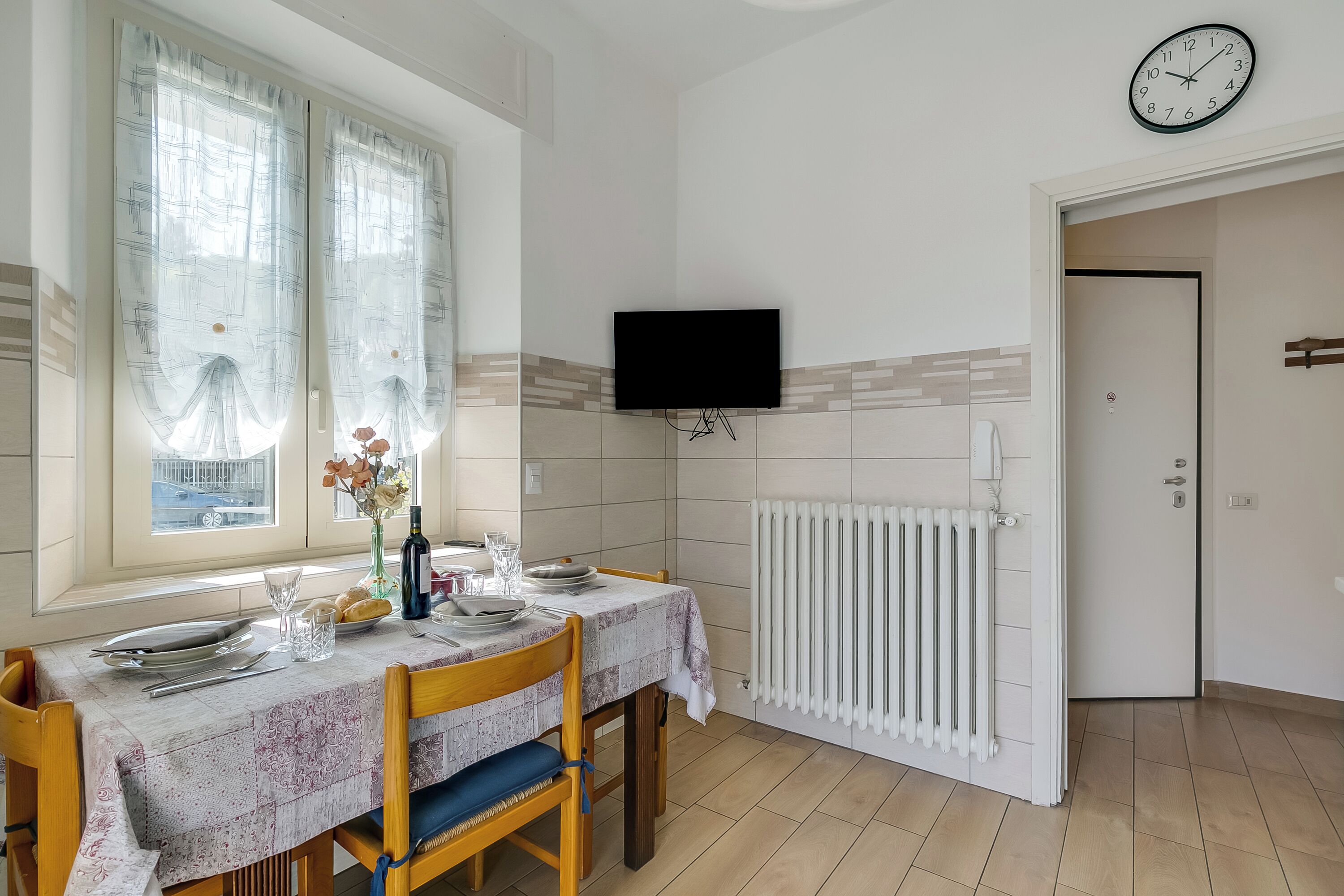 Un coin repas avec une table dressée pour deux personnes, une fenêtre avec des rideaux, un téléviseur fixé au mur et un radiateur.