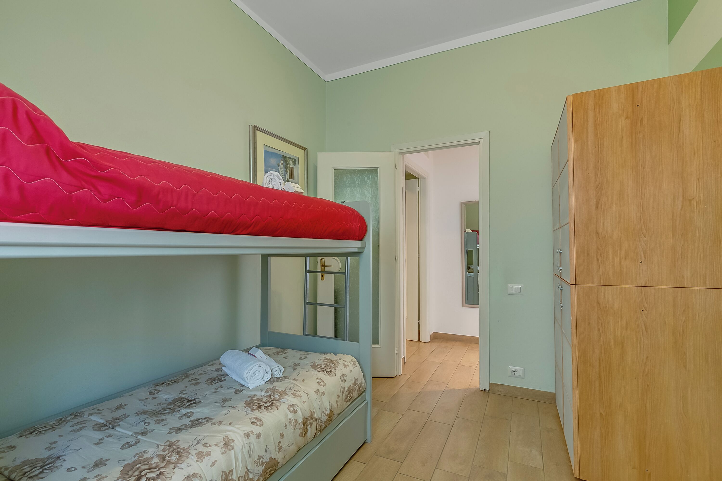 Ein Etagenbett mit rotem oberen Bett und einem unteren Bett mit floraler Bettdecke. Dazu ein Holzschrank rechts und eine Tür zu einem anderen Raum.