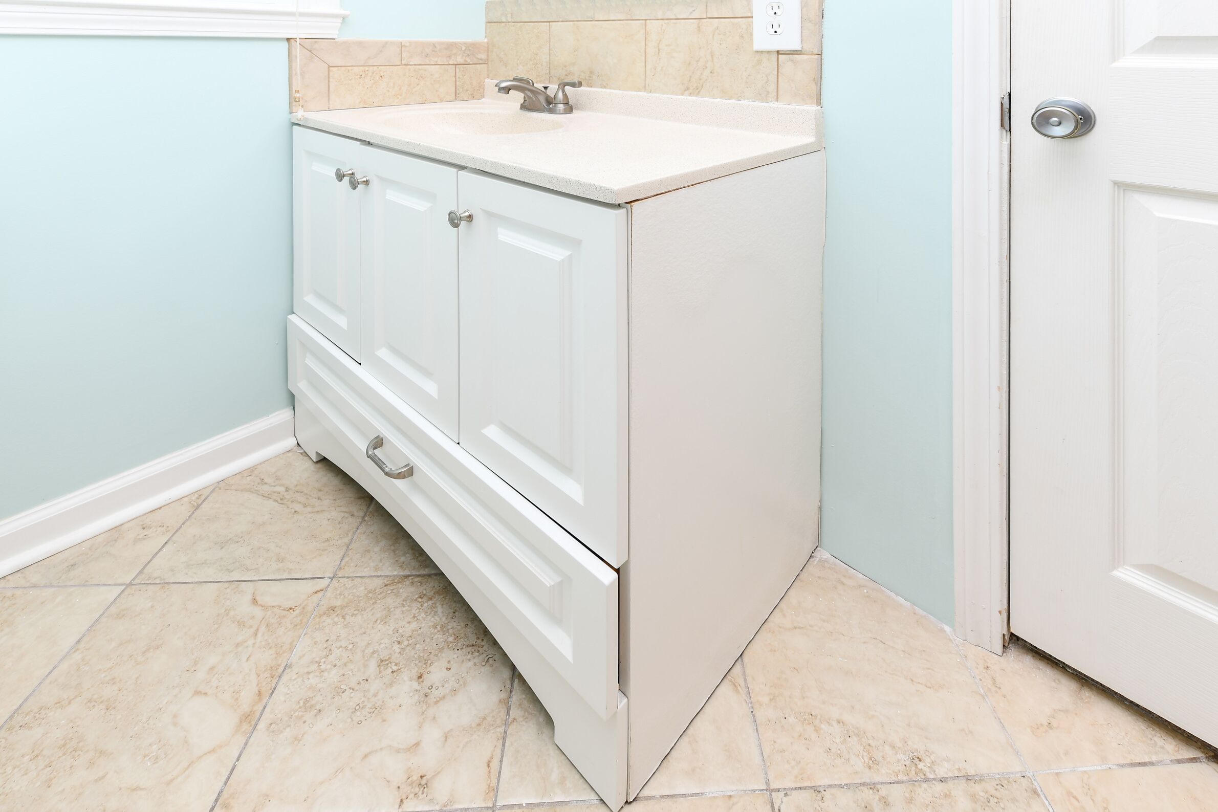 Une petite salle de bain blanche, avec un lavabo et un meuble, un sol carrelé et une porte.