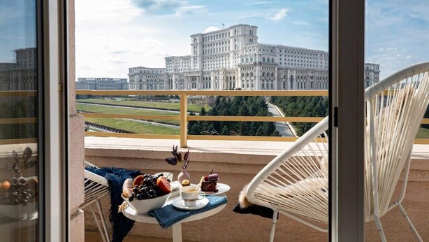 A balcony with a table set for breakfast, a white chair, and a view of a large building. - Qult·co - Monarch (București)