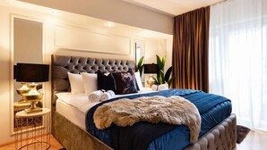 A bedroom with a tufted headboard, a large bed with a blue bedspread and a fur throw, a bedside table with a lamp, and a large mirror on the wall. - Qult·co - Monarch (București)