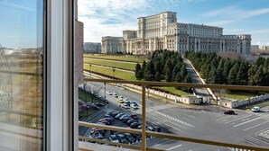 A view of a large, historic building with a symmetrical facade and multiple windows, seen from a balcony overlooking a parking area and a road. - Qult·co - Monarch (București)