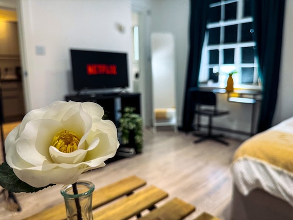 A room with a bed, a desk, a chair, a TV displaying Netflix, and a white flower in a glass vase.
