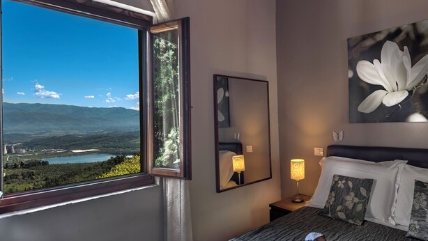 A hotel room with a large window offering a scenic view of a lake and mountains, a neatly made bed with striped bedding, a bedside lamp, and a framed artwork of a flower.