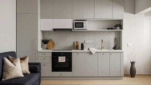 Una cocina moderna con armarios grises, un microondas y un lavavajillas. Hay un sofá oscuro con cojines y un jarrón con plantas secas.