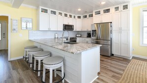 A kitchen with white cabinets, a stainless steel refrigerator, a microwave, and a sink. There are three stools at the island. - Oceanfront 4 BR Beach House with a Private Pool! Surfin' Turf (Surfside Beach)
