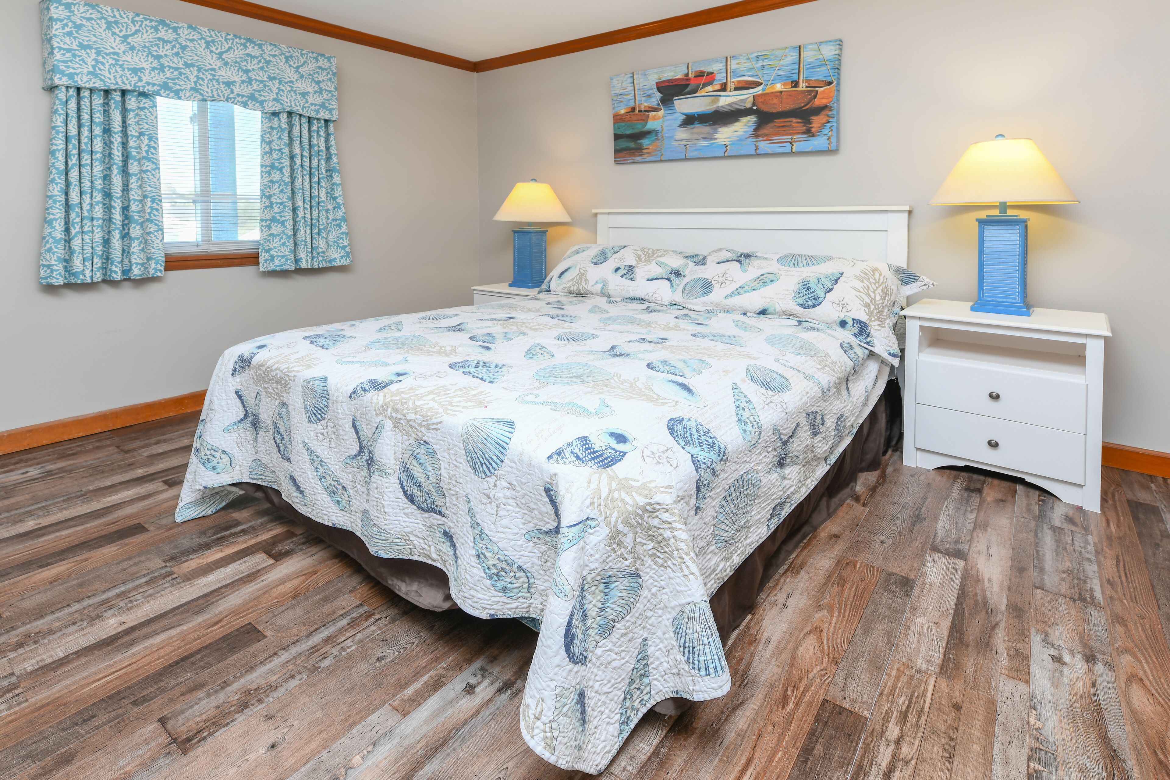 A bedroom with a bed, wooden flooring, a window with curtains, two bedside tables with lamps, and a painting on the wall.