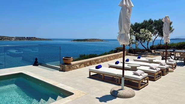 Ein Poolbereich mit Liegestühlen und Sonnenschirmen mit Blick auf ein klares blaues Meer und entfernte Landstriche.