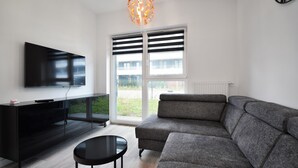 Una sala de estar moderna con televisor de pantalla plana, mueble de entretenimiento negro, sofá seccional gris, una mesa de centro redonda y un ventanal con persianas.