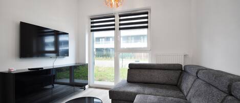 Una sala de estar moderna con televisor de pantalla plana, mueble de entretenimiento negro, sofá seccional gris, una mesa de centro redonda y un ventanal con persianas.