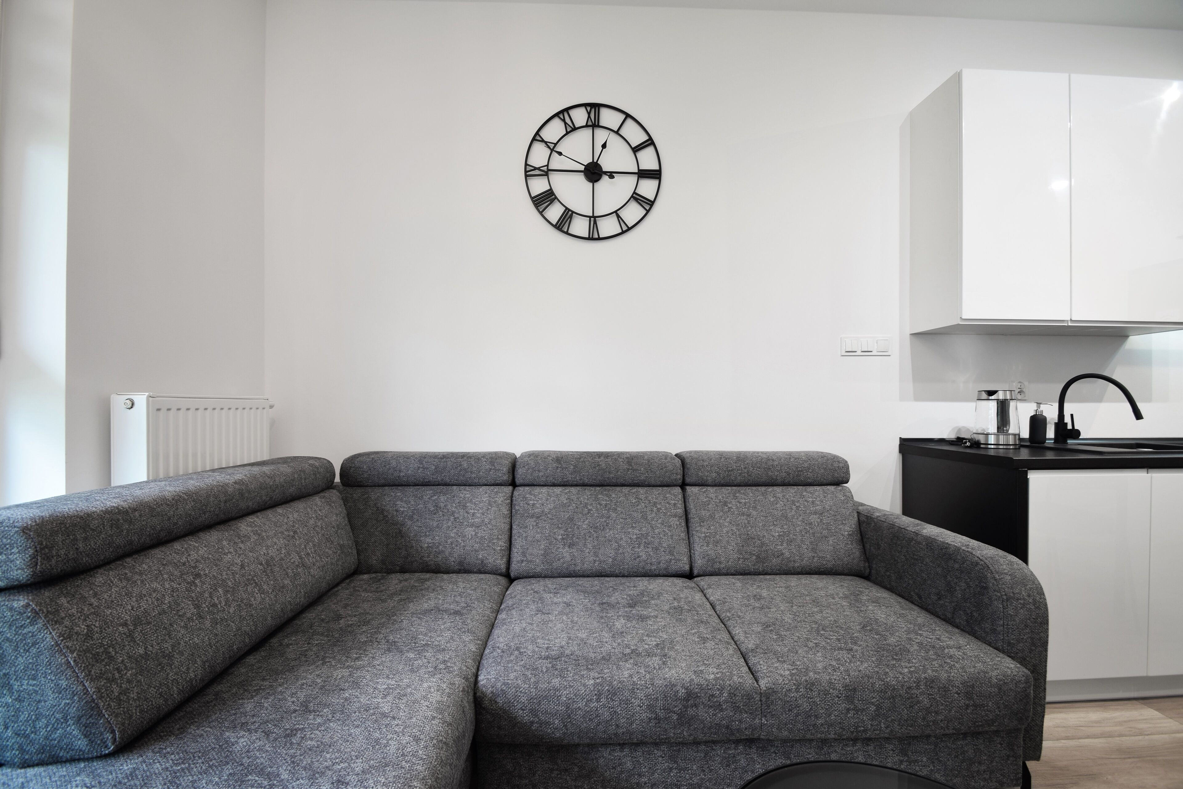 A modern living room with a grey sectional sofa, a wall-mounted clock, and a kitchen area with white cabinets and a black countertop.
