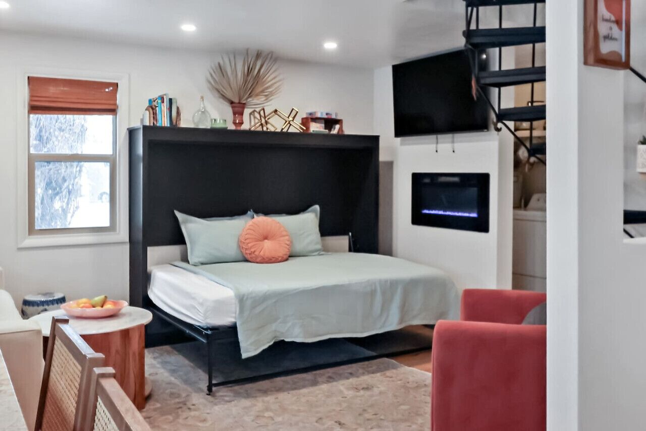 A modern bedroom with a bed, a TV mounted on the wall, a small table with a bowl of fruit, and a chair.