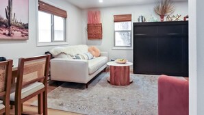 A living room with a sofa, a coffee table, and a TV. There are two windows with blinds, a cactus painting on the wall, and a bookshelf with books and decorative items. - Cozy Hideaway Retreat Unit 4 near Durango, CO. (Durango)