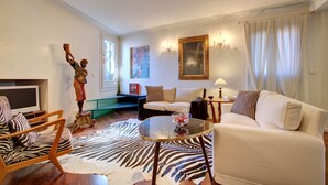 Una sala de estar con un sofá, un sillón, una mesa de centro de vidrio y un tapete estampado de cebra. Se observa una estatua, un cuadro y una lámpara sobre una mesita auxiliar.