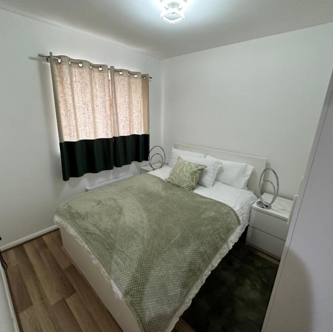 Ein Schlafzimmer mit Bett, Nachttisch, Lampe, einem Fenster mit Vorhängen und einer Deckenleuchte.