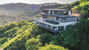 A modern multi-story house with large glass windows, situated on a hillside surrounded by lush greenery. - Modern 6 BR Ocean View Villa with pingpong table, daily breakfast & cleaning (Playa Tamarindo)