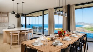 A modern dining area with a wooden table set for six, large windows offering a view of the ocean, and a kitchen with a round island and bar stools. - Modern 6 BR Ocean View Villa with pingpong table, daily breakfast & cleaning (Playa Tamarindo)