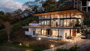 Une maison moderne à deux étages, dotée de grandes fenêtres en verre, d’une terrasse en bois et d’une allée.