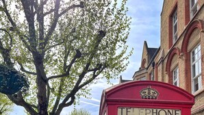 A red telephone booth with a crown emblem, located on a tree-lined street with brick buildings.