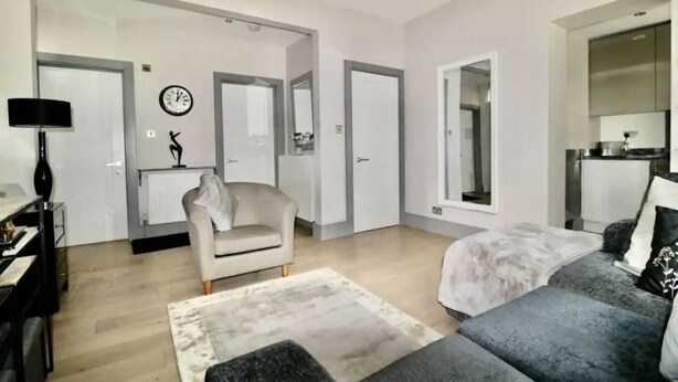 A modern living room with a sofa, a chair, a rug, and a kitchen area visible through an open door.