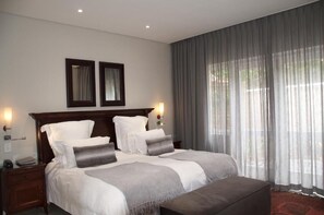 A modern hotel room with a large bed, two bedside tables, a nightstand, a window with sheer curtains, and a view of the outdoors.