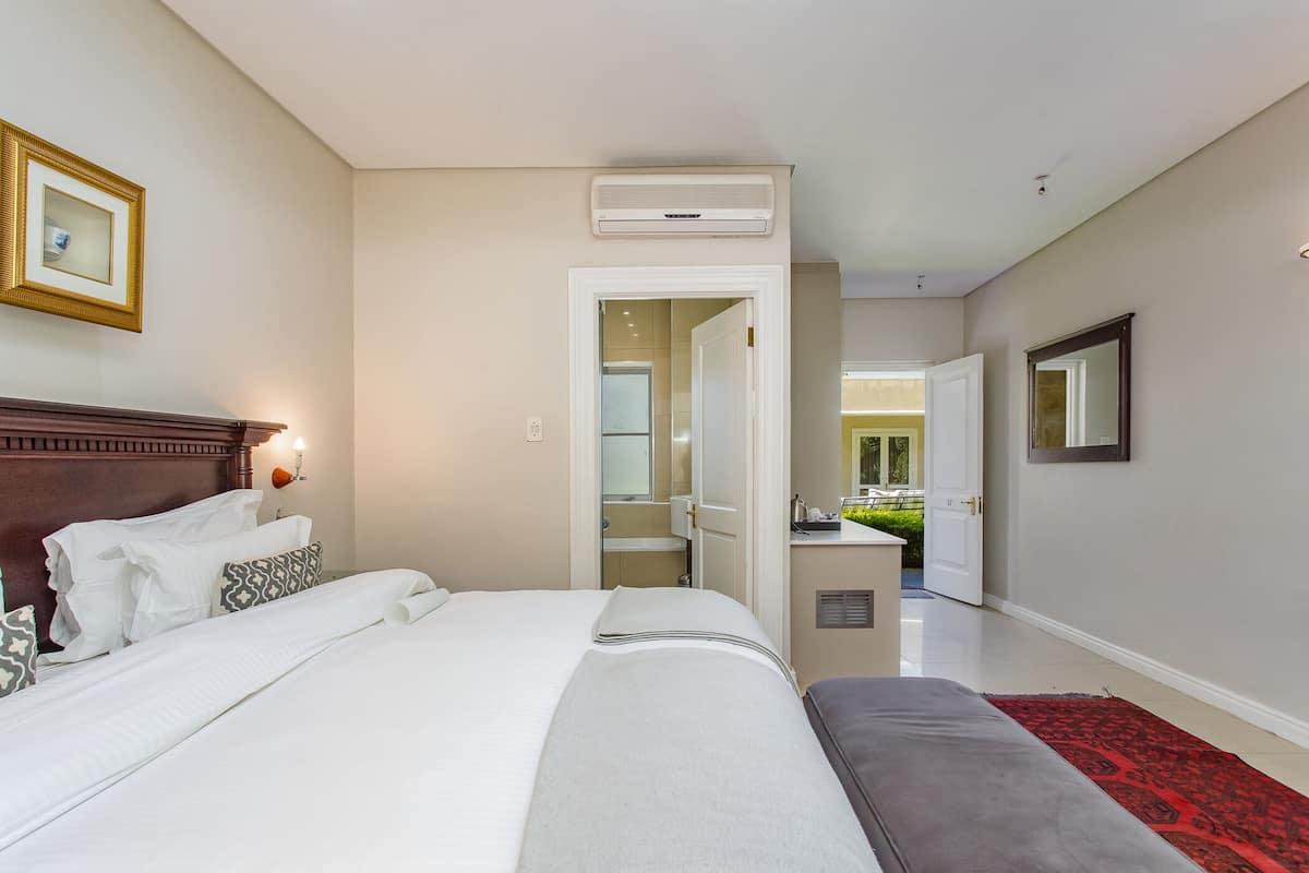 A hotel room with a neatly made bed, a bedside table with a lamp, a large mirror, and a door leading to a bathroom and a small kitchenette.