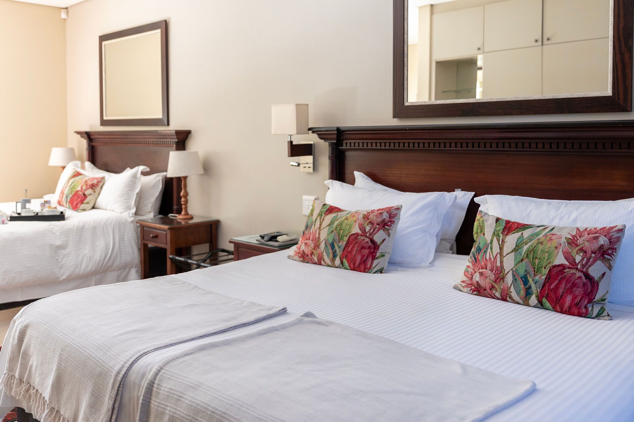 A hotel room with two beds, each with white linens and floral-patterned pillows. There is a wooden headboard, a nightstand with a lamp, and a mirror on the wall.