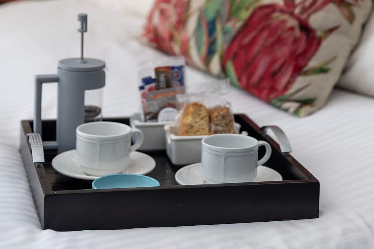 A breakfast tray with a coffee maker, cups, and pastries on a bed with a floral pillow.