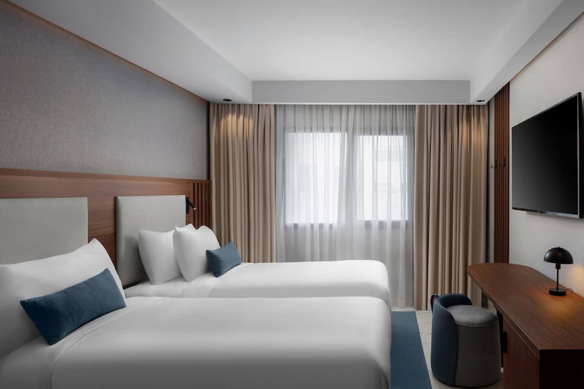 A hotel room with two beds, a desk, a chair, a TV, and a window with curtains.
