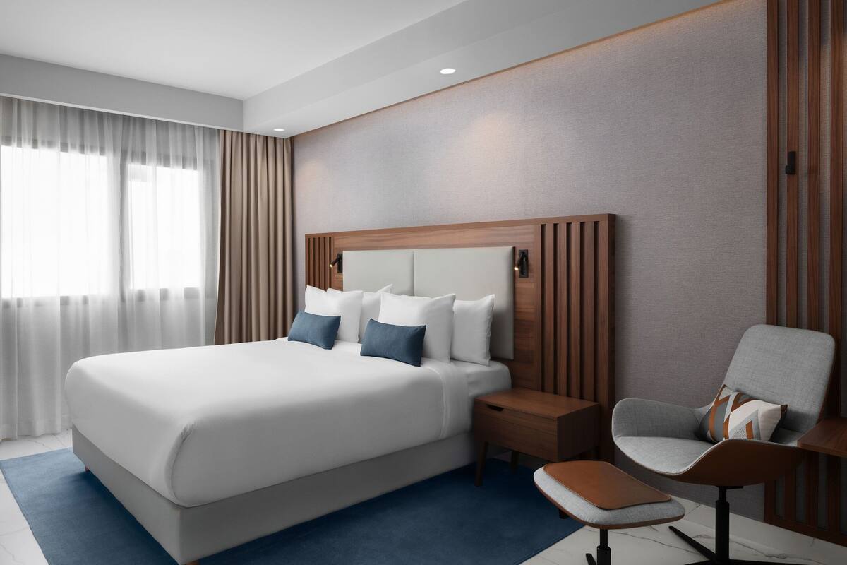 A modern hotel room with a large bed, a wooden headboard, a small table, a chair, and a window with curtains.