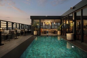 A rooftop pool area with seating and a dining area, surrounded by glass walls and potted plants.