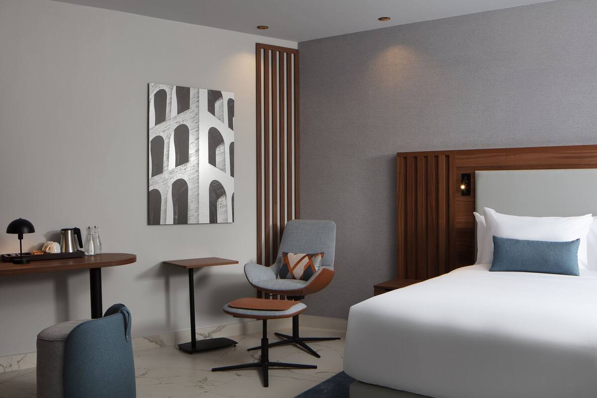 A modern hotel room with a large bed, a seating area with a chair and ottoman, a small table with a lamp, and a wall-mounted artwork.