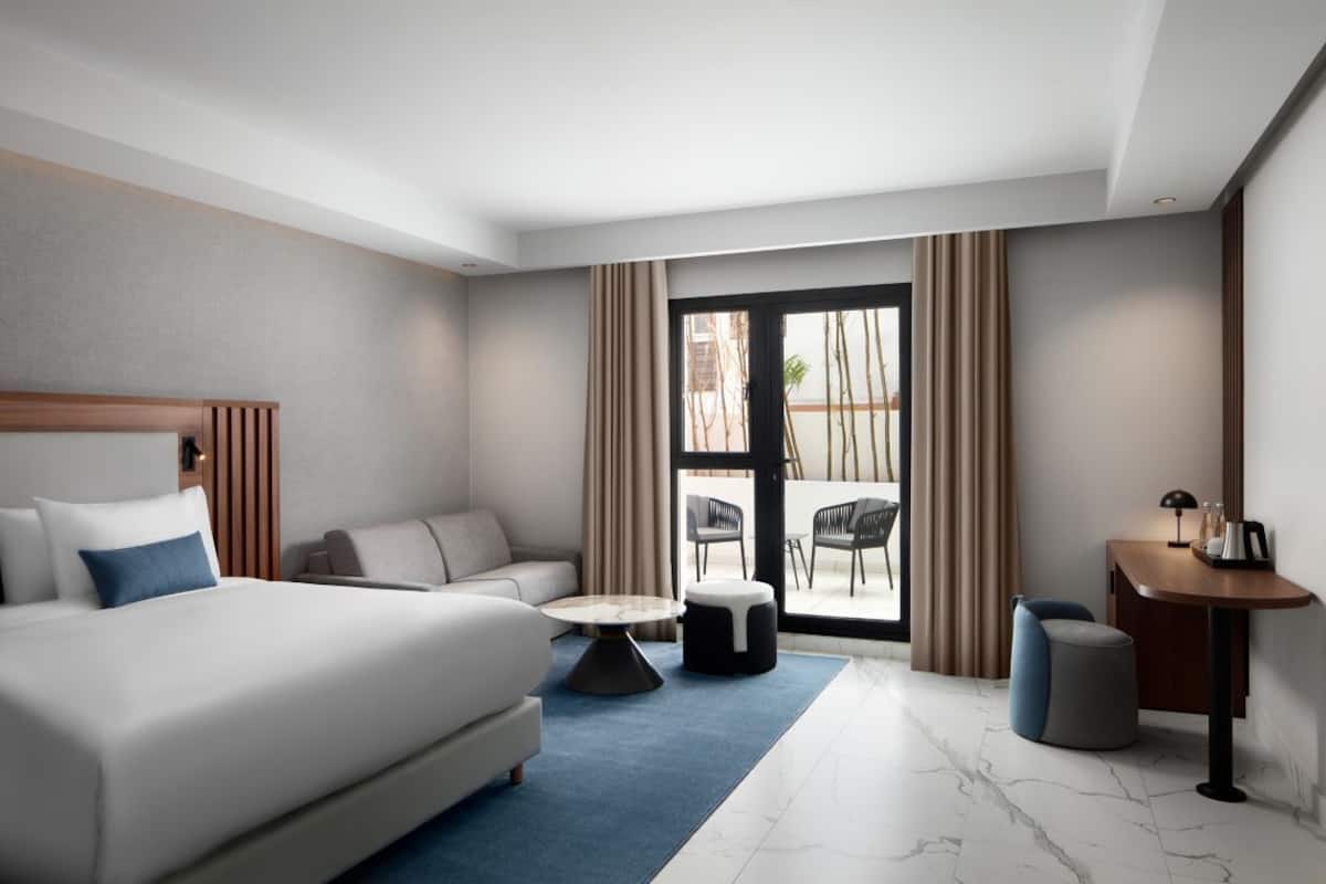 A hotel room with a bed, sofa, round coffee table, ottoman, and a desk with a lamp.