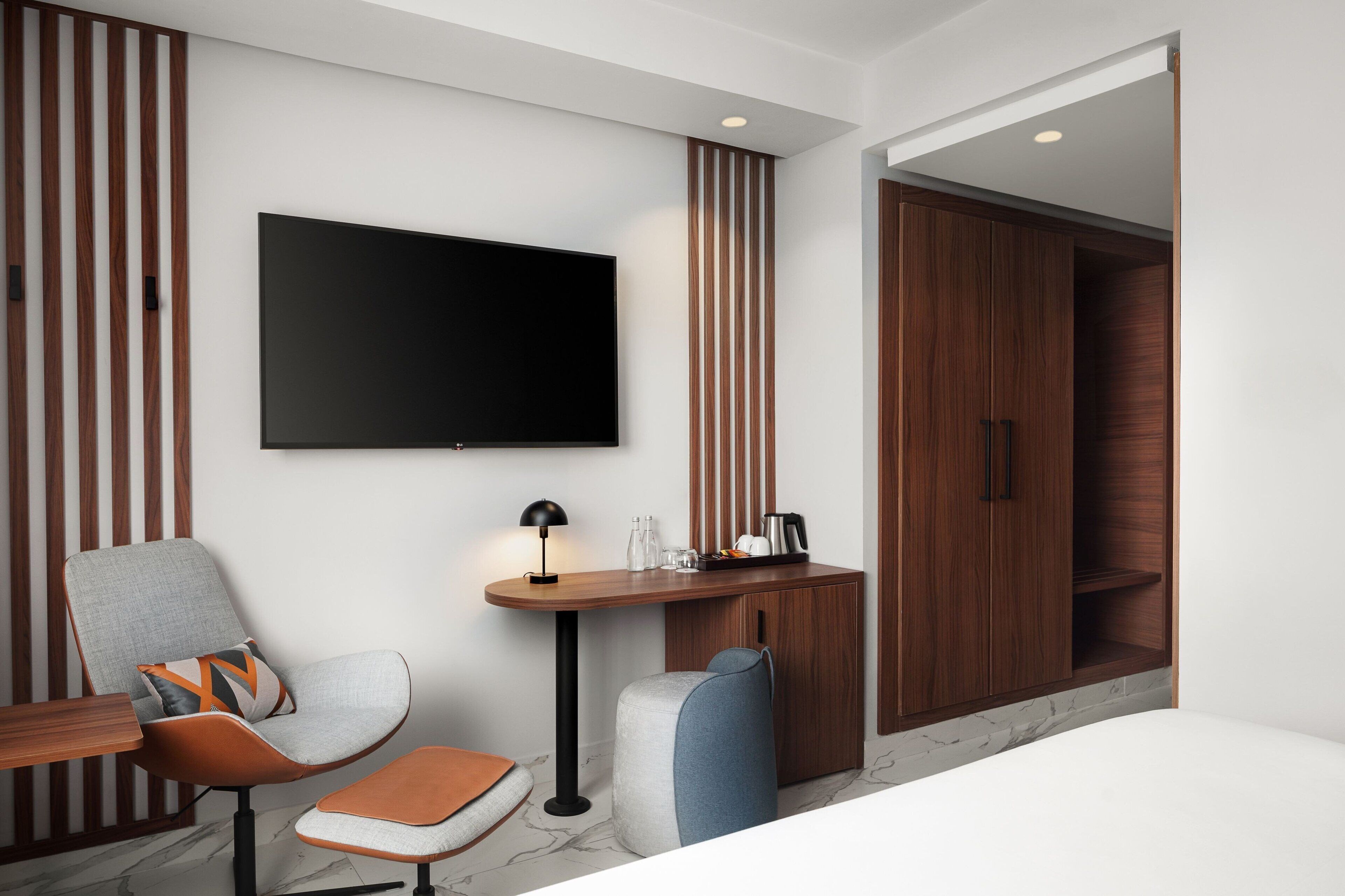 A modern hotel room with a flat-screen TV, a wooden wardrobe, a desk with a lamp, and a comfortable seating area.