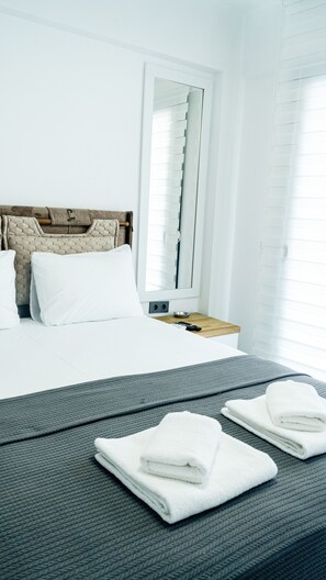 A hotel room with a bed, a bedside table with a bowl and remote control, and a window with blinds. - Port Marmaris Rooms (Marmaris)