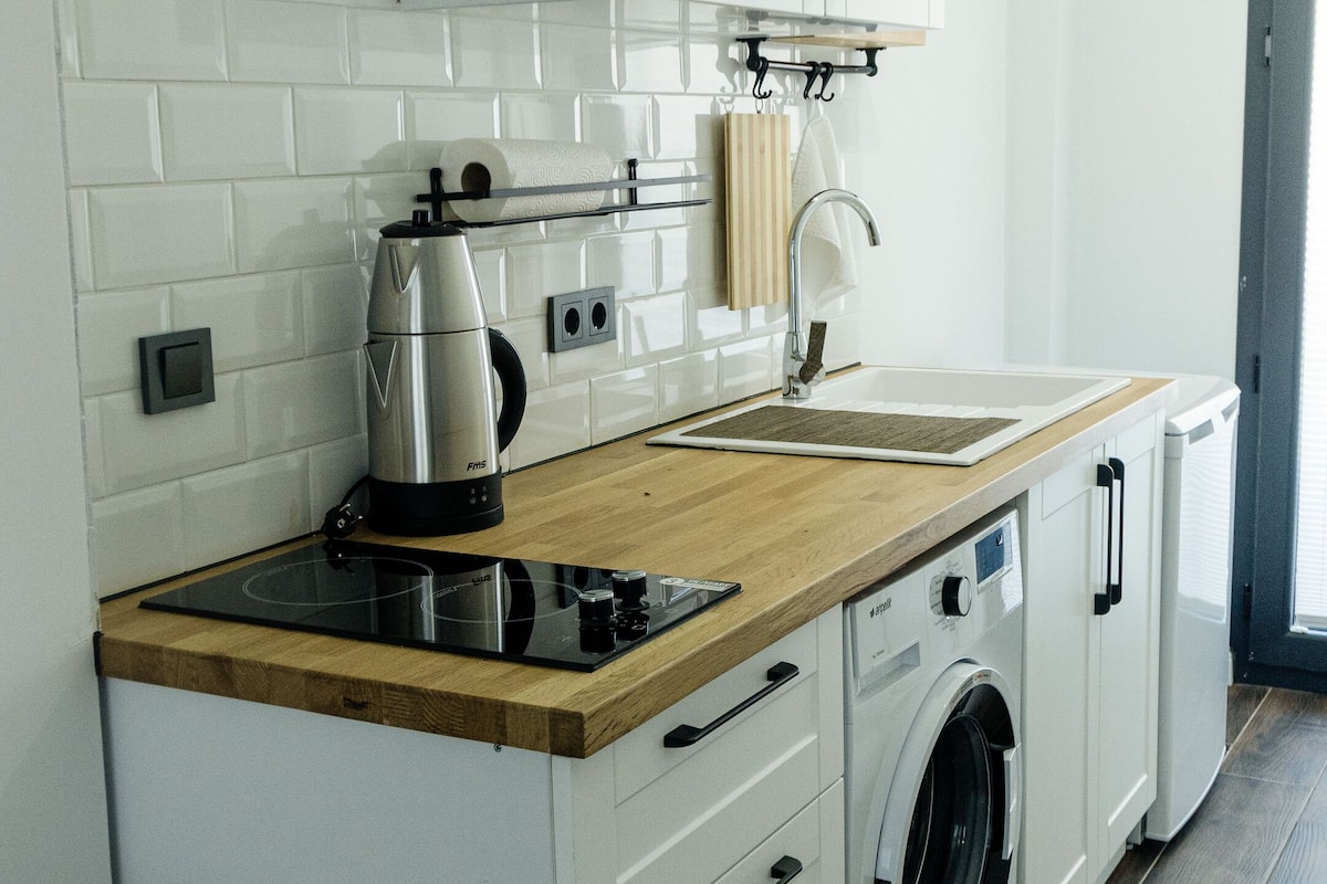 Een kleine keuken met witte kastjes, een houten aanrechtblad, een ingebouwde wasmachine en een hanglamp.