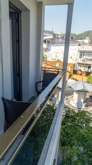 A balcony with a metal railing, a black cushioned chair, and a view of a street with buildings and a sign for 'My Capella'. - Port Marmaris Rooms (Marmaris)